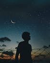 A lone silhouette stands beneath a crescent moon and starry sky, evoking a sense of solitude and wonder.